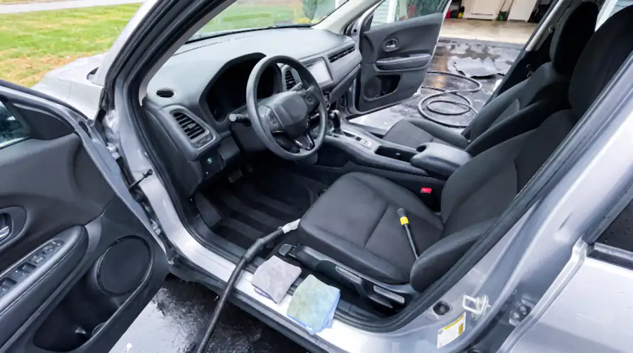 Illustration of the same cabin after an interior detail: cloth clean, fresh vacuum lines in the carpet, dashboard matte and dust-free, cup holders empty. The steam wand and extractor hose run out of the open door and the floor mats are drying outside on the wet driveway.