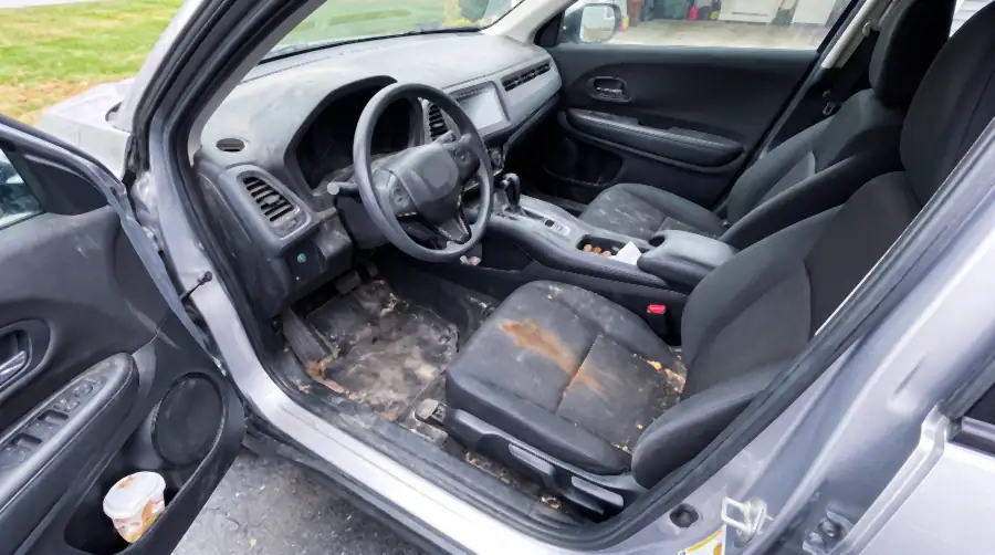 Illustration of the same crossover's cabin before an interior detail: dried mud and salt caked on the driver's floor mat, sand ground into the carpet, a coffee stain on the seat cushion, dust film over the dashboard, litter in the door pocket.