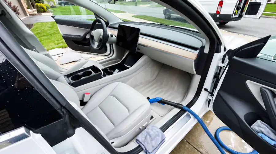 Illustration of the same cabin after an interior detail: white upholstery genuinely white again, pale carpet even in colour with fresh vacuum lines, wood strip and flat dash dust-free, screen clean. The steam wand rests on the sill and mats dry outside on the wet driveway.