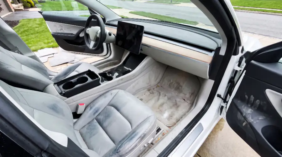Illustration of the same EV's cabin before an interior detail: white seat bolsters greyed and streaked with denim dye transfer, dirty handprints down the white door card, pale footwell carpet stained with mud and salt, smears across the blank touchscreen.