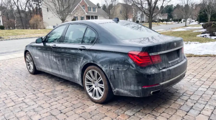 Illustration of a black luxury saloon on a brick driveway before a detail: a chalky grey-white film of road salt over the whole body killing all reflection, salt streaks down the lower doors, wheels grey with brake dust.