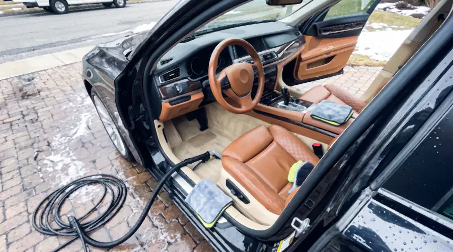 Illustration of the same cabin after an interior detail: tan leather evenly clean and satin, seams free of grit, pale carpet uniform with fresh vacuum lines, wood trim deep and dust-free, mats drying outside on the wet bricks.