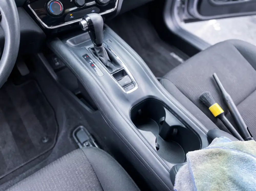 Illustration of the same console after cleaning: cup holders empty and clean to the bottom of the wells, every seam brushed out, gloss black trim spotless, with a soft brush and a blower nozzle lying beside it.