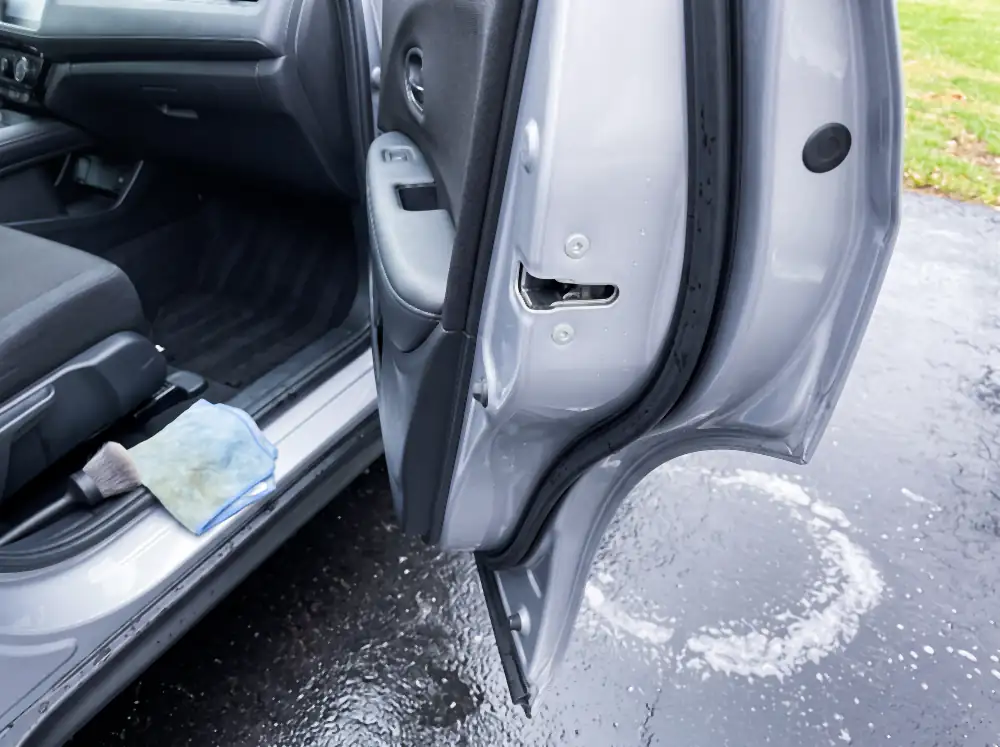 Illustration of the same door jamb after cleaning: painted jamb bright and clean into the corners, weather seal deep black with fresh water droplets on it, striker clean bare metal, with a damp towel and brush on the sill and the wet driveway below.