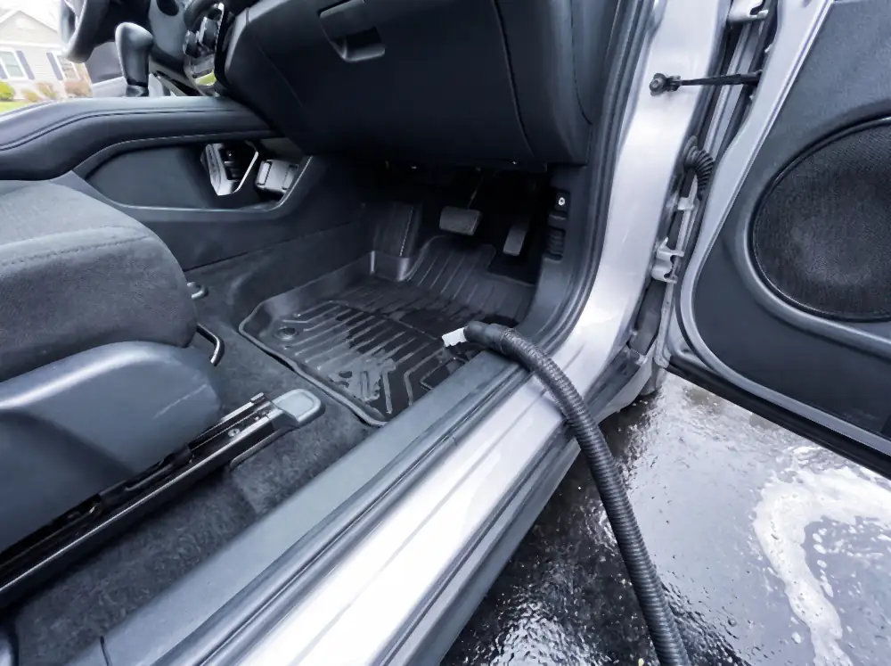 Illustration of the same footwell after cleaning: the rubber mat scrubbed and laid back in wet and deep black, the carpet uniformly dark with fresh vacuum lines, with the extractor hose across the sill and a second mat drying outside on the wet driveway.