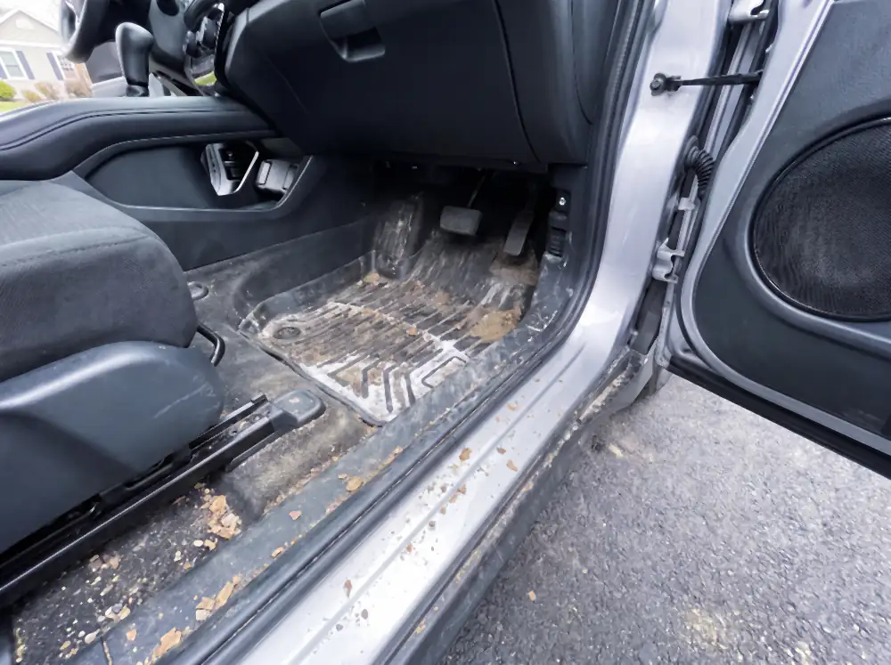 Illustration of a driver's footwell before cleaning: the rubber mat heaped with dried mud, sand and a thick crust of white road salt in every channel, the carpet around it grey-brown and matted with grit.