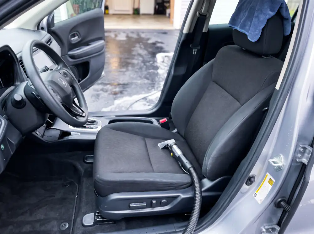 Illustration of the same seat after cleaning: cloth evenly clean and uniformly dark, seams clear of grit, fabric still slightly damp from extraction, with the extractor nozzle resting on it and a damp towel over the backrest.