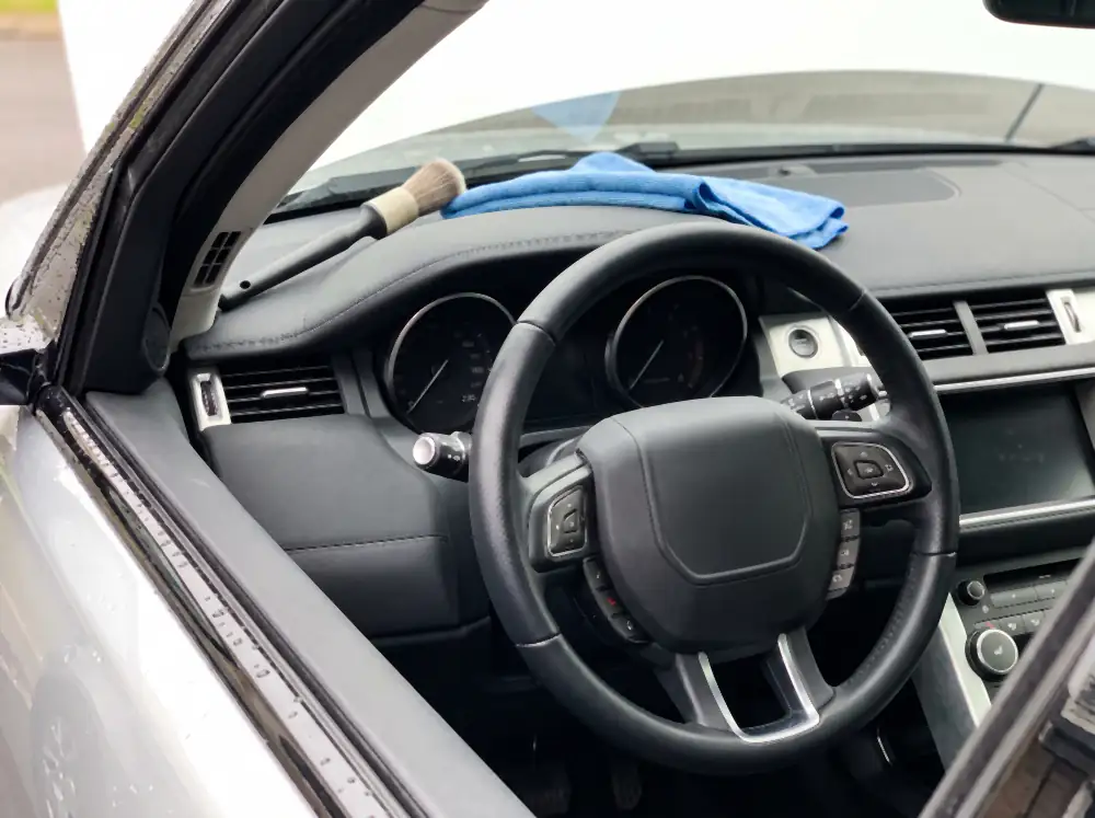 Illustration of the same steering wheel after cleaning: the leather rim matte and evenly clean with no shine, perforations clear, cluster glass spotless, dashboard satin and dust-free, with a detailing brush and damp towel resting on the cluster hood.