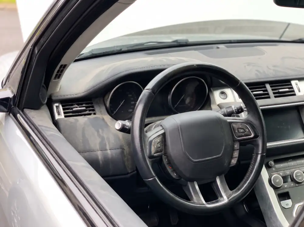 Illustration of a steering wheel before cleaning: a dark greasy shine polished into the leather at the ten and two positions, dirt in the perforations, a thick film of dust across the dashboard top with finger streaks through it.