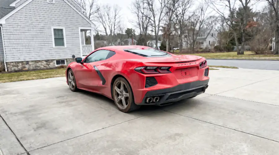Illustration of a red mid-engine sports coupe on a concrete driveway before a detail: a dull grey-brown layer of dust and winter grime over the whole body, dried spots down the rear deck, brake dust on the wheels.
