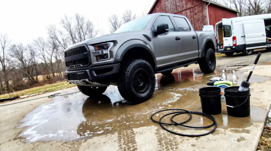 Illustration of the same pickup after a detail: paint evenly clean and matte, flares deep black, tyre sidewalls clean and dressed, wheels spotless. The concrete pad is soaked dark with wet tread prints, foam is running off toward the gravel and the hose, buckets and open van are in shot.