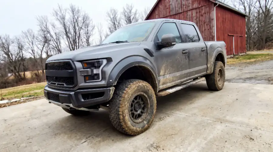 Illustration of a lifted grey crew-cab pickup on a concrete pad before a detail: dried mud sprayed up the flanks and tailgate, a crust of white salt along the rockers, mud packed into the wheels and knobby tyre treads, splattered glass.