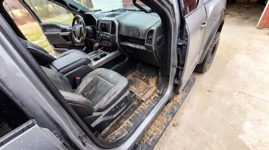 Illustration of the same pickup's cab before an interior detail: dried mud clods and gravel heaped across the rubber floor mat, mud smeared up the door card and side step, grit in the leather seams, dust over the dashboard.