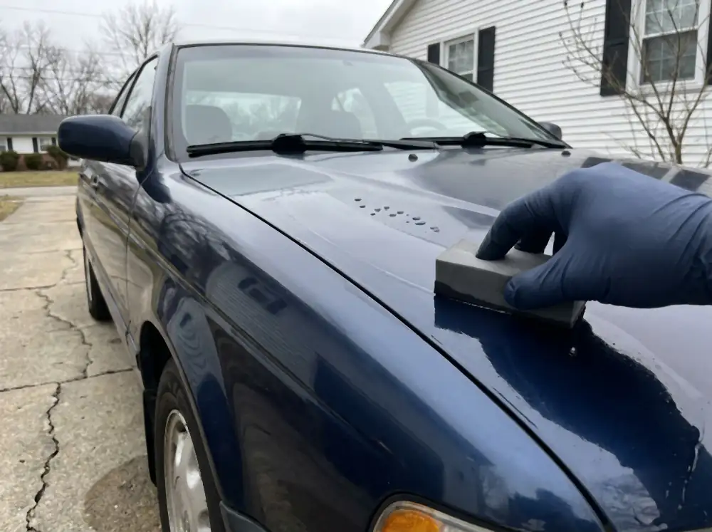 A gloved hand drawing a coating applicator block across the bonnet of a navy car, leaving a visibly slicker trail behind it