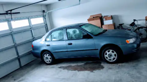 A car parked inside a home garage with heavy condensation beaded across its windows and bodywork, and a damp patch on the concrete floor beneath it.