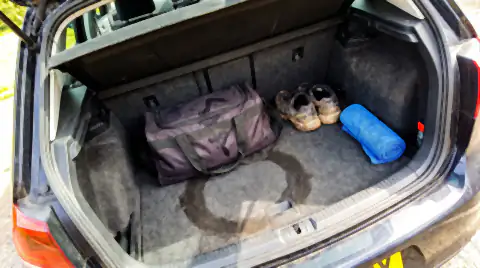 A damp gym bag, trainers and a rolled towel in an open car boot, with a dark stain ring on the carpet where the bag has been sitting.