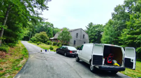 A long asphalt driveway curving away from a New England house, with a car parked halfway down it and a service van behind, doors open.