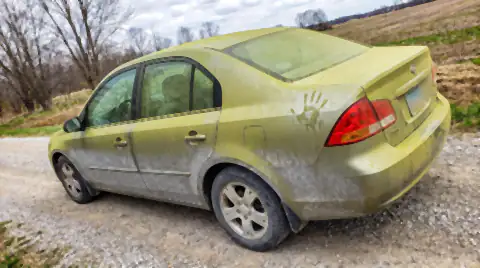 A car on a rural gravel road covered in a thick yellow-green film of tree pollen, with pale gravel dust coating the lower panels.