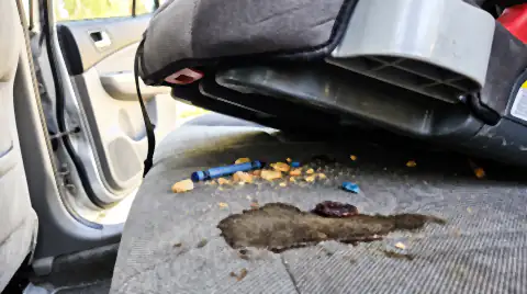 A child seat lifted out of a car's back seat, revealing crushed crumbs, a broken crayon and a stained patch of upholstery underneath.