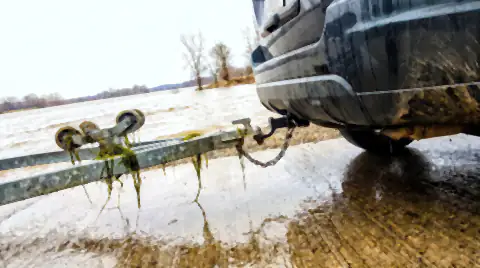 The rear of a car with an empty boat trailer hitched to it at a river boat ramp, river water and weed still on the trailer rollers.