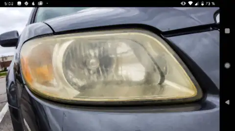 A car headlight whose plastic lens has gone cloudy, yellowed and crazed with oxidation, next to the clean painted wing beside it.