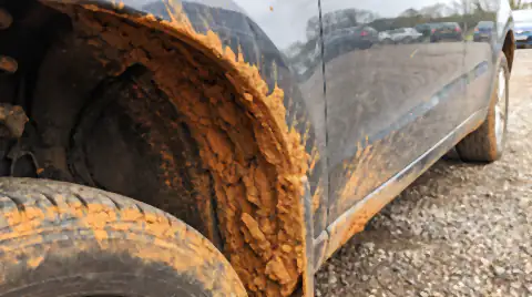 A car wheel arch and rocker panel caked in thick orange-brown clay farm mud, drying and cracking into plates.