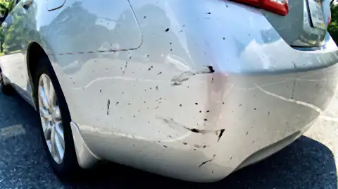 Small black tar spots splattered across the lower rear quarter of a light-coloured car, some smeared where they have been wiped.