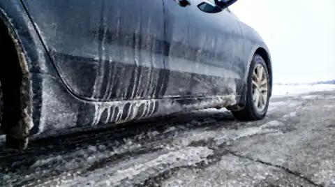 A car's rocker panel and lower door crusted with dried white road-salt brine streaks, with salt-stained slush on the road alongside.