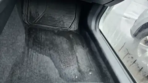 A car footwell with a dark wet patch spreading through the carpet from under the mat, and condensation fogging the window above it.