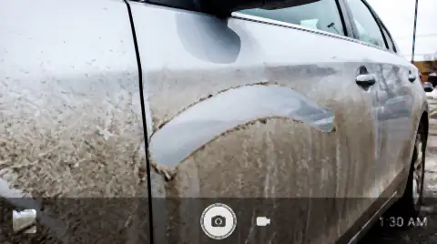 The flank of a silver car coated in grey-brown highway road film, with one clean finger-swipe through it showing the paint underneath.