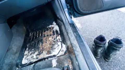 A car footwell in ski season with melting snow, car-park grit and a white salt crust on the rubber mat, and ski boots on the ground outside.