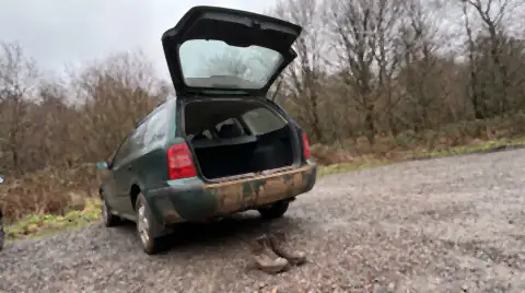 The open boot sill and rear bumper of a car at a woodland trailhead smeared with drying trail mud, muddy boots on the gravel below.