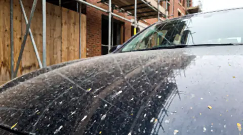 A dark car bonnet beside a building site, coated in fine grey construction dust with hard speckles of dried overspray stuck to it.