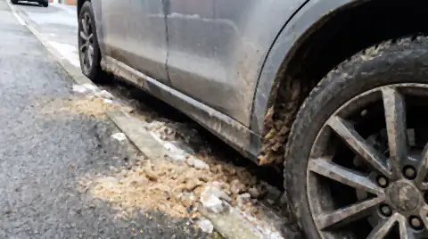 The lower body and wheel of a car on a steep hill street coated in gritty brown winter road sand, with loose grit piled at the kerb.
