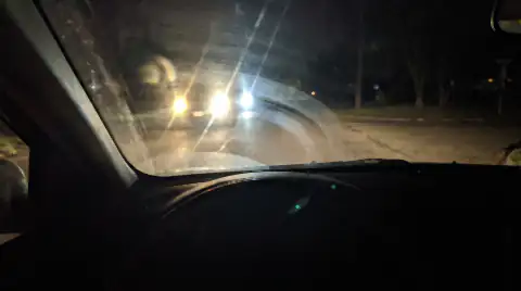 The view out through a car windscreen at night, a hazy film on the inside of the glass scattering oncoming headlights into starburst glare.
