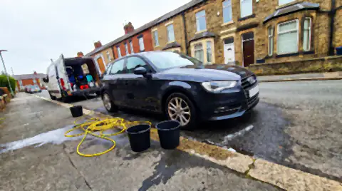 A car being detailed at the kerb on a residential street with no driveway, hose and buckets on the pavement and a service van in front.
