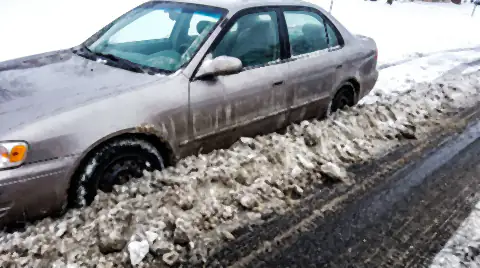 A street-parked car with a heavy ridge of dirty grey snow and frozen slush thrown up its flank by a passing plough.