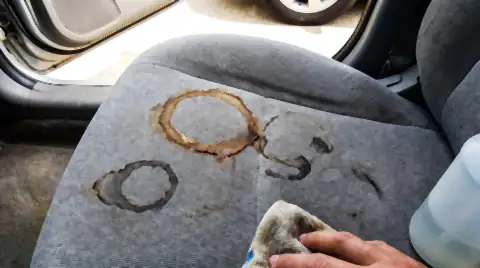 A grey cloth car seat with a dried coffee stain and a sticky soda ring, both showing brown tide-marks at their edges.