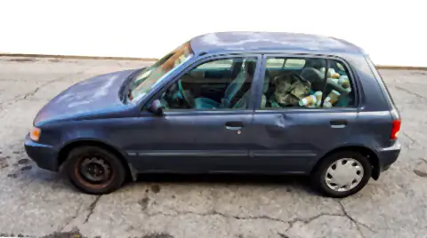 An older, well-used hatchback parked on a city street: faded roof paint, one mismatched wheel and a cluttered back seat.