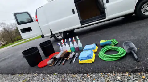 A mobile detailer's kit laid out on a driveway: buckets with grit guards, brushes, unlabelled spray bottles, folded microfibre towels and a coiled hose.