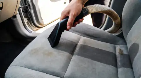 A hot-water extractor nozzle drawn across a cloth car seat, leaving a clean wet band behind it and dirty brown water in the recovery hose.