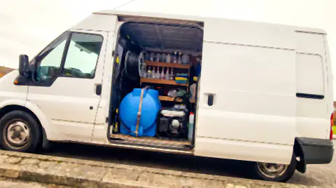 The open rear doors of a plain white mobile detailing van showing the kit inside: a strapped-down water tank, generator, pressure washer and shelves of bottles.